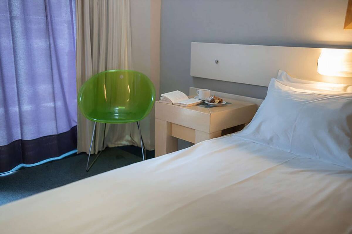 A hotel room with a bed, a green chair, a bedside table with a phone and a cup, and a window with curtains.