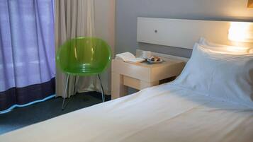 A hotel room with a bed, a green chair, a bedside table with a phone and a cup, and a window with curtains.
