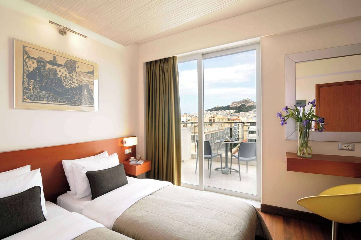 A hotel room with two beds, a balcony with a table and chairs, a large window, and a painting on the wall.