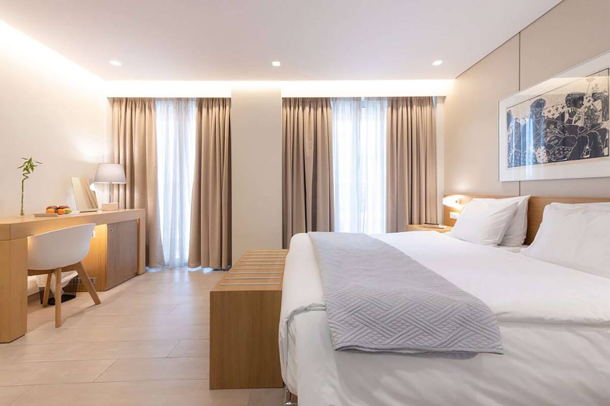 A modern hotel room with a bed, desk, and chair, large windows with curtains, and a painting on the wall.