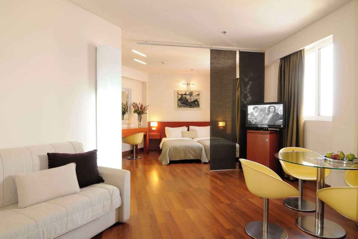 A hotel room with a sofa, a bed, a dining table with chairs, a TV, and a wall-mounted artwork.