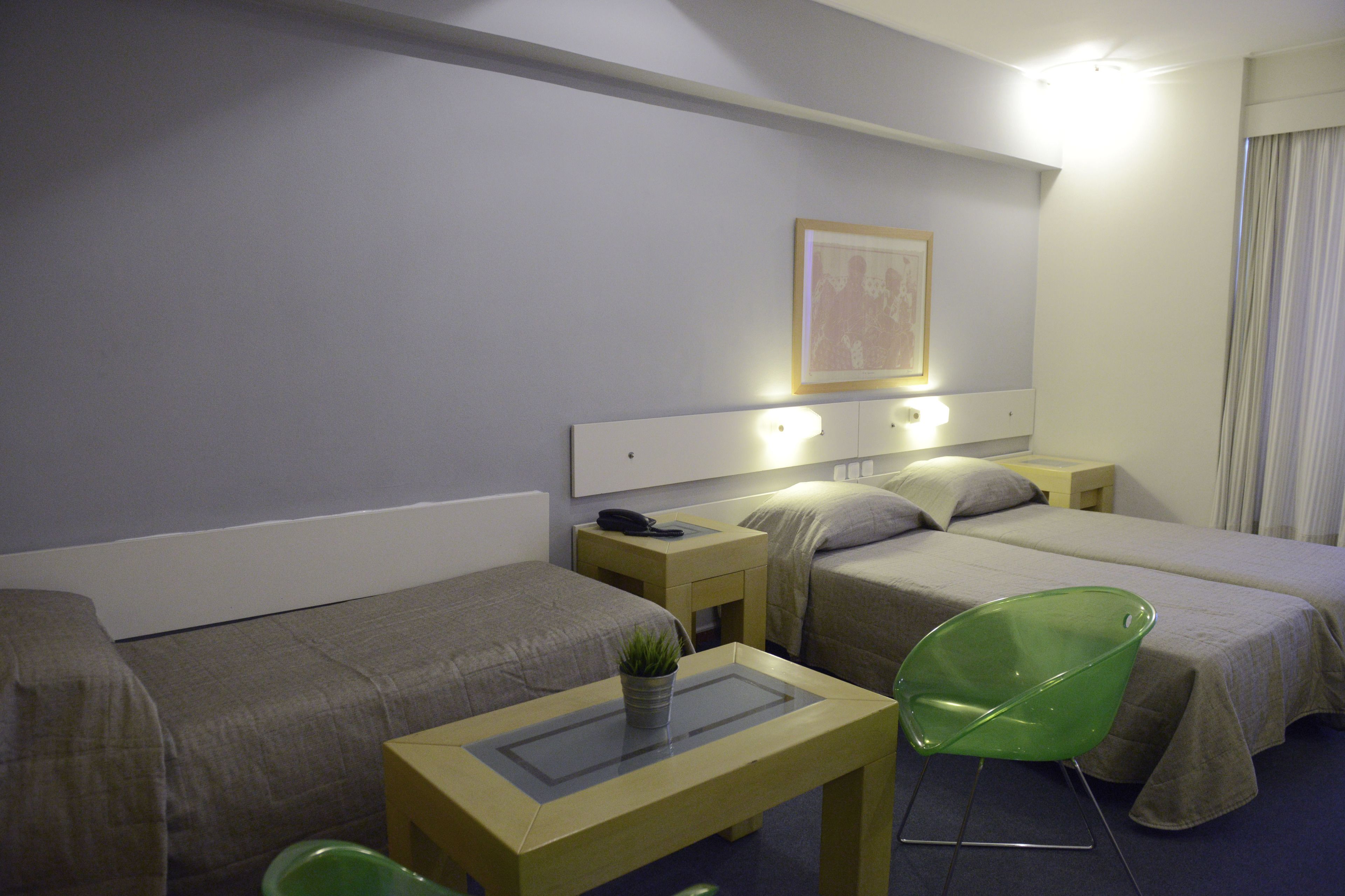 A hotel room with two single beds, a small table with a plant, a green chair, and a bedside table with a lamp.