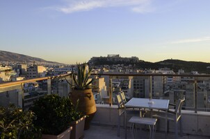 Varanda com mesa e cadeiras, com vista para um horizonte urbano que inclui uma colina e um edifício semelhante à Acrópole.