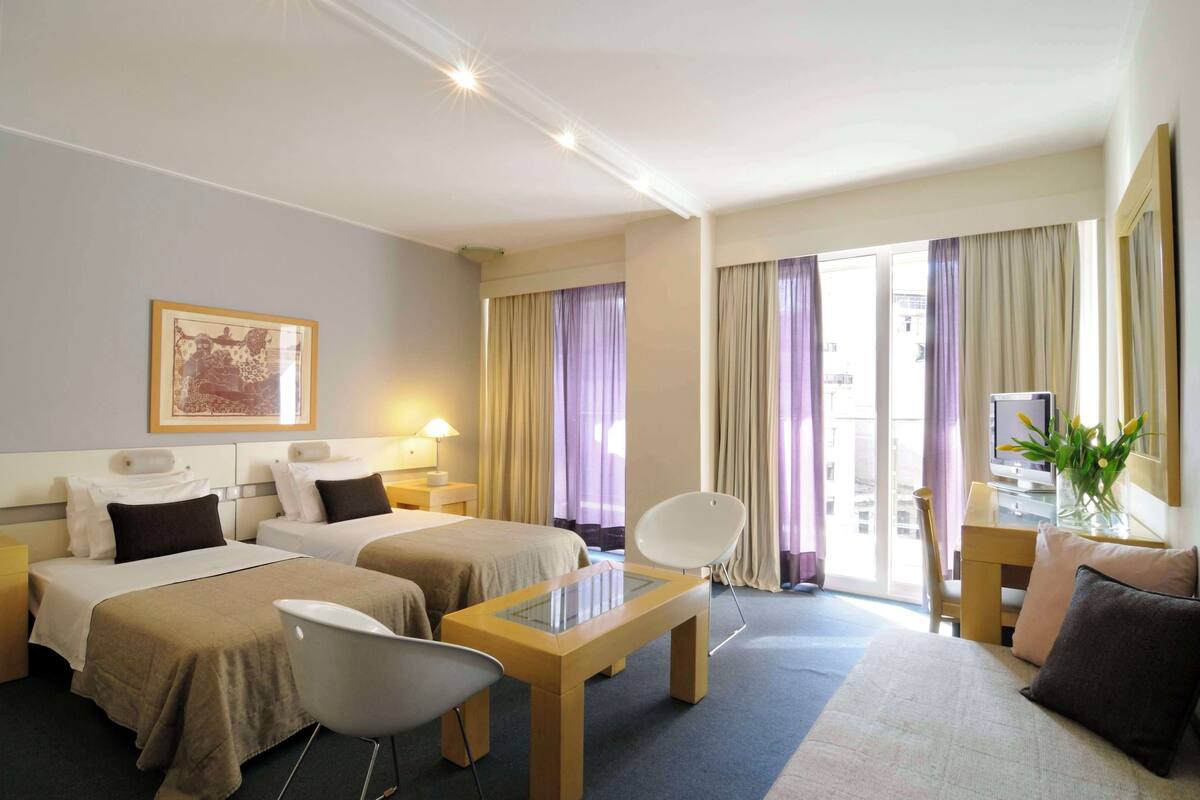 A hotel room with two beds, a desk, a chair, a TV, and a window with curtains.
