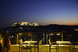 Μια ταράτσα με τραπέζια και καρέκλες, από την οποία διακρίνεται ένα ιστορικό κτίριο φωτισμένο σε ένα φόντο ηλιοβασιλέματος.