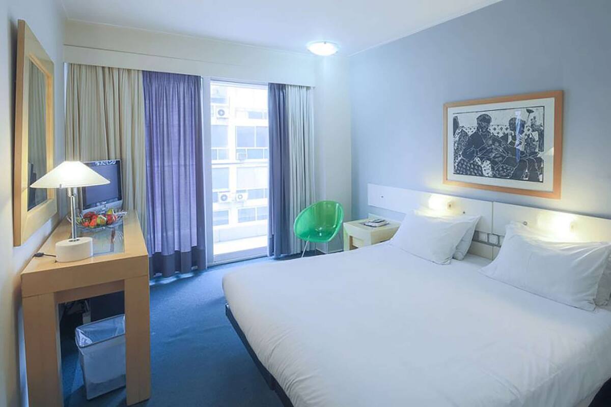 A hotel room with a large bed, a desk with a lamp, a chair, a small table, and a framed picture on the wall.