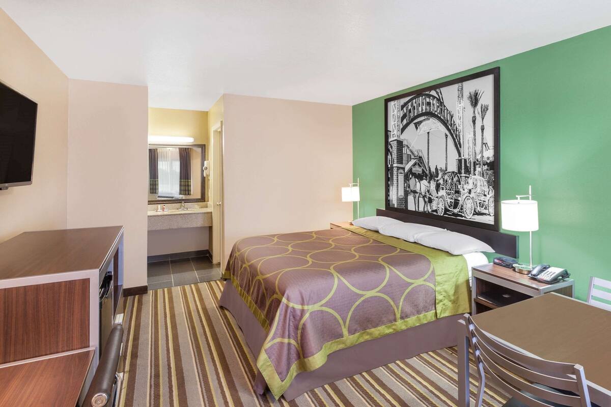 A hotel room with a bed, a desk, a chair, a television, a bathroom with a mirror, and a wall-mounted picture.