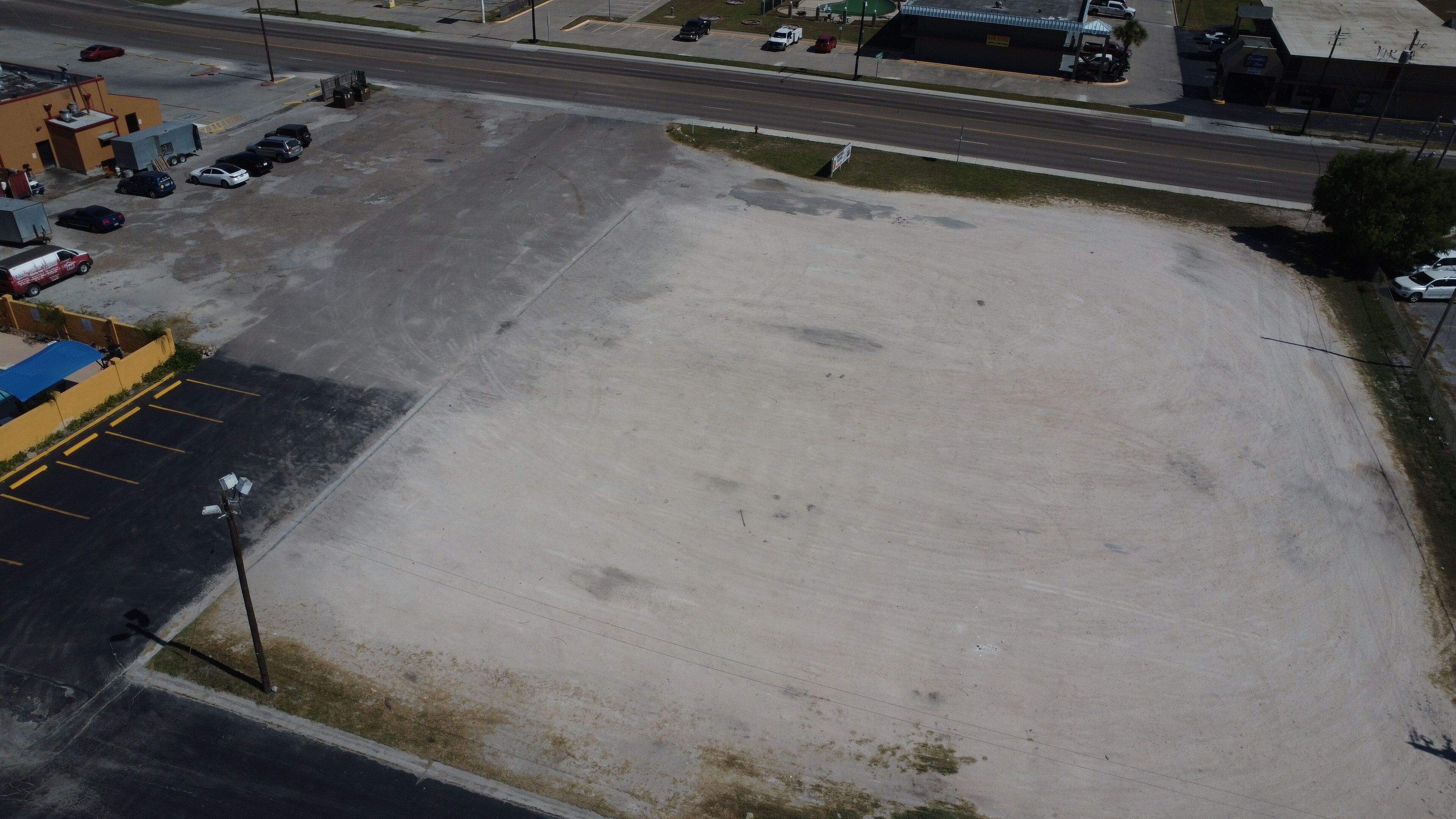 Un estacionamiento vacío junto a una carretera, con algunos coches aparcados y un edificio al fondo.