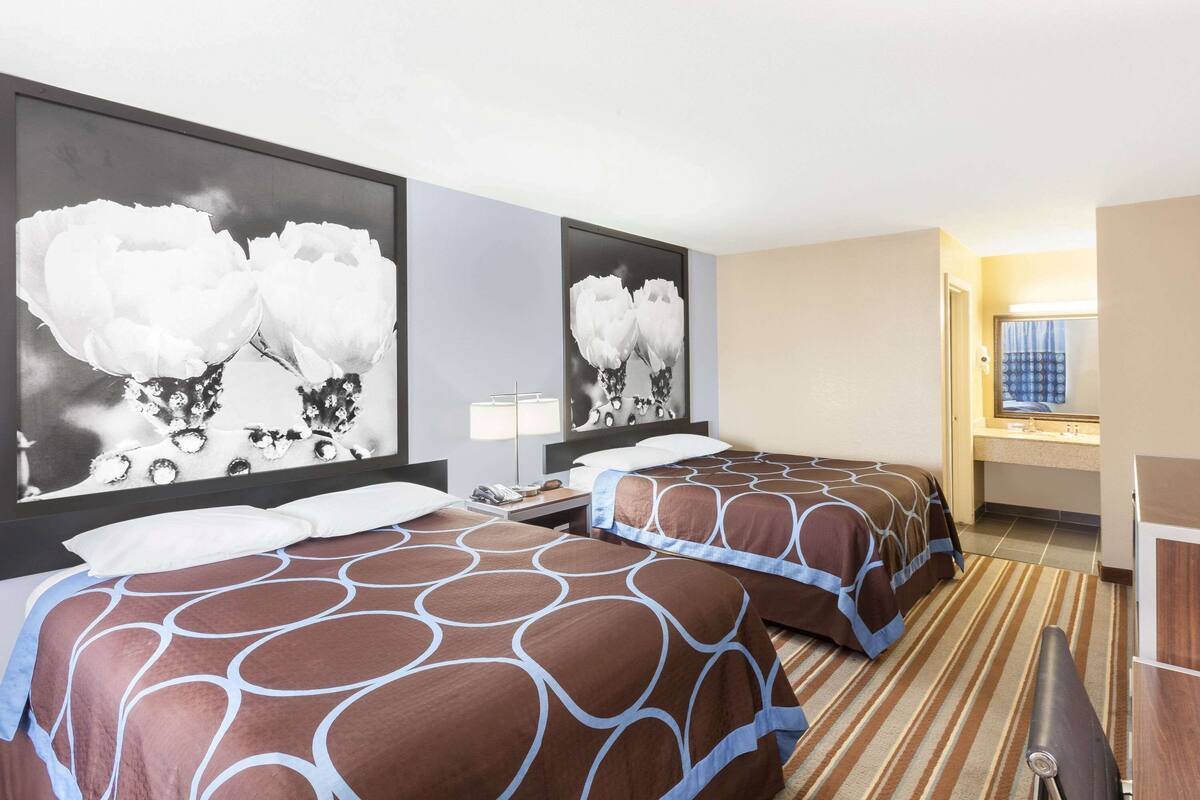 A hotel room with two beds, a large wall art of flowers, a bedside table with a lamp, and a bathroom visible through an open door.