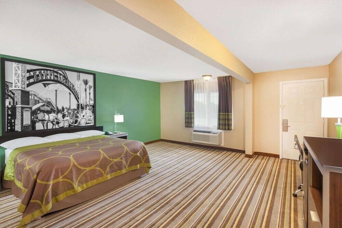 A hotel room with a bed, a desk, a window with curtains, an air conditioning unit, and a wall-mounted picture.