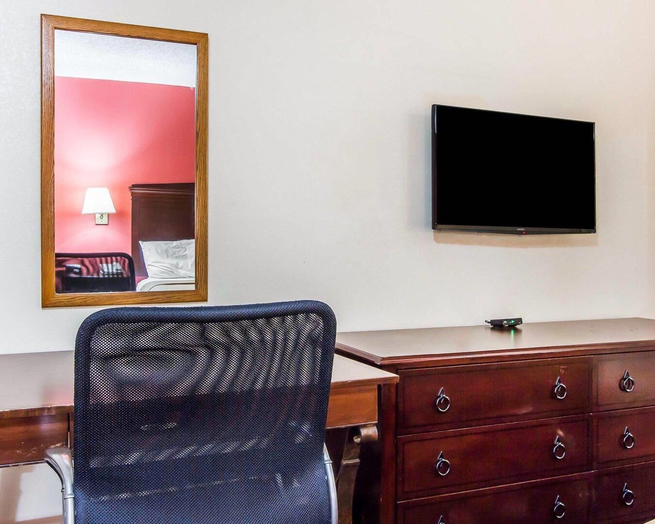 A hotel room with a wooden dresser, a television mounted on the wall, a chair, and a mirror reflecting part of the room.