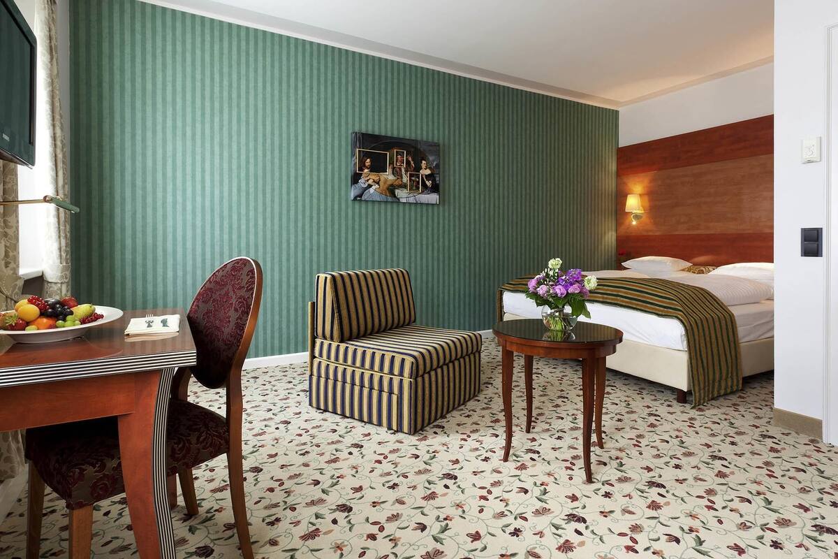 Ein Hotelzimmer mit Bett, Stuhl, kleinem Tisch mit Vase, Esstisch mit Obst und einem Bild an der Wand.