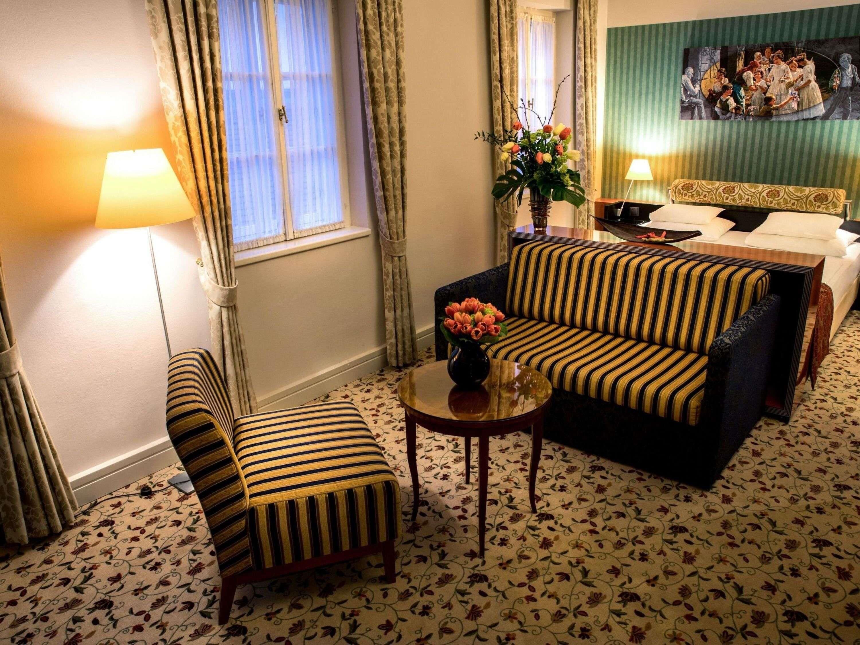 A hotel room with a sofa, armchair, bedside table, and a painting on the wall.