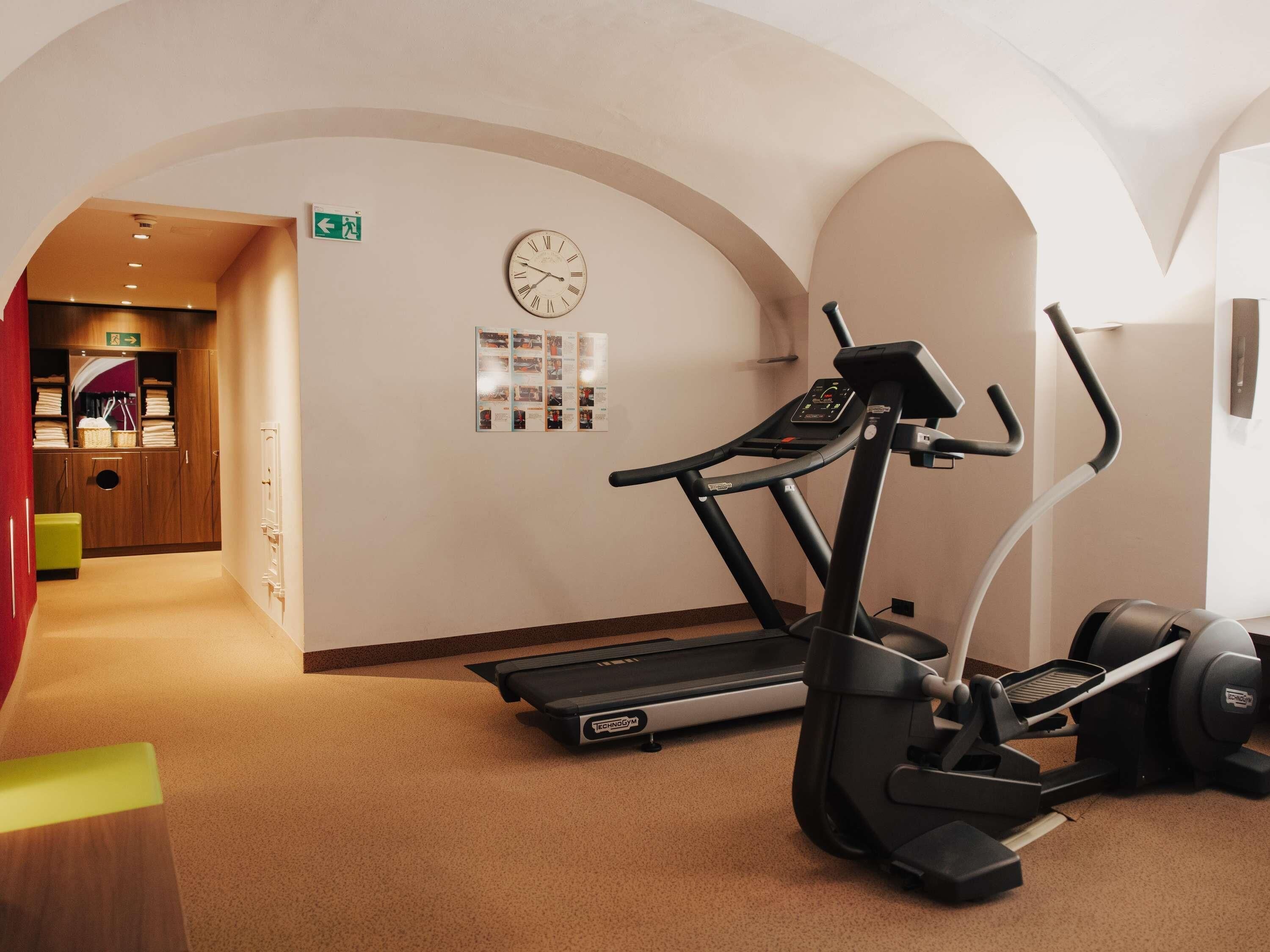 A fitness area with a treadmill, elliptical, and stationary bike, a clock, and a bulletin board with flyers.