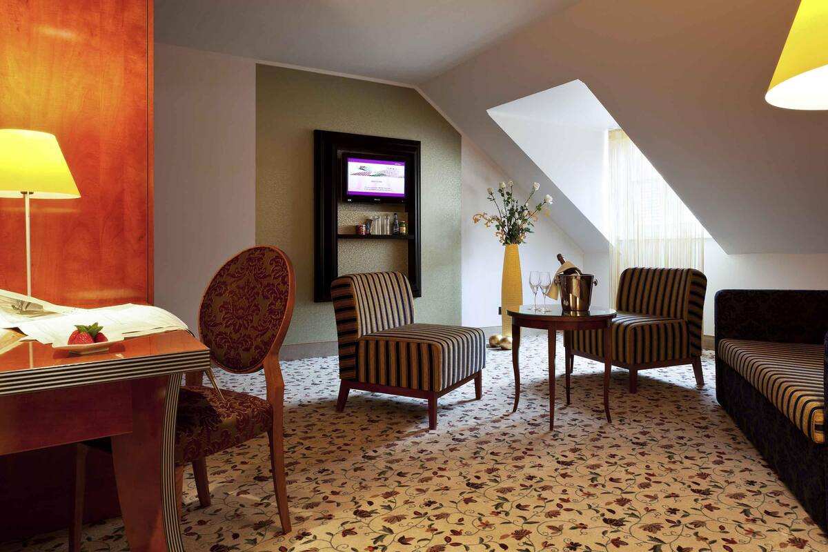 Ein Hotelzimmer mit Esstisch, zwei Sesseln, einer Couch, einem an der Wand befestigten Fernseher, einem kleinen Tisch mit einer Vase und einer Champagnerflasche sowie einem gemusterten Teppich.