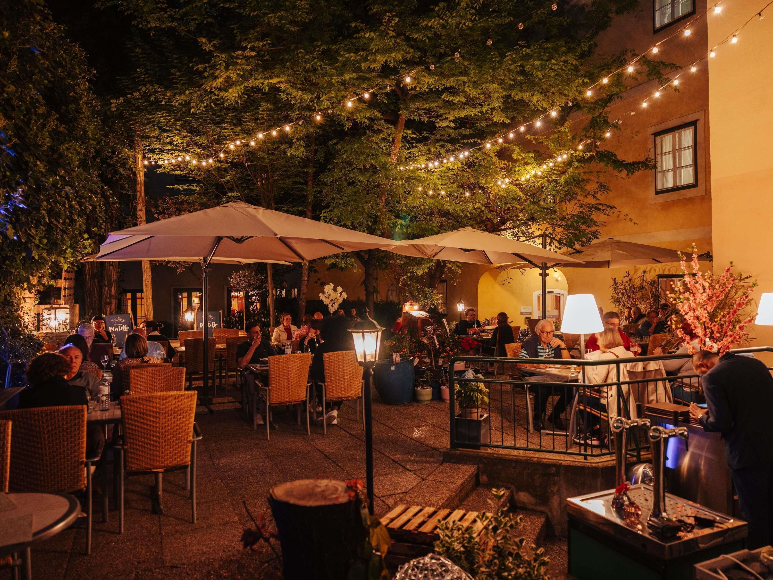 An outdoor dining area with wicker chairs and tables, string lights, and umbrellas.