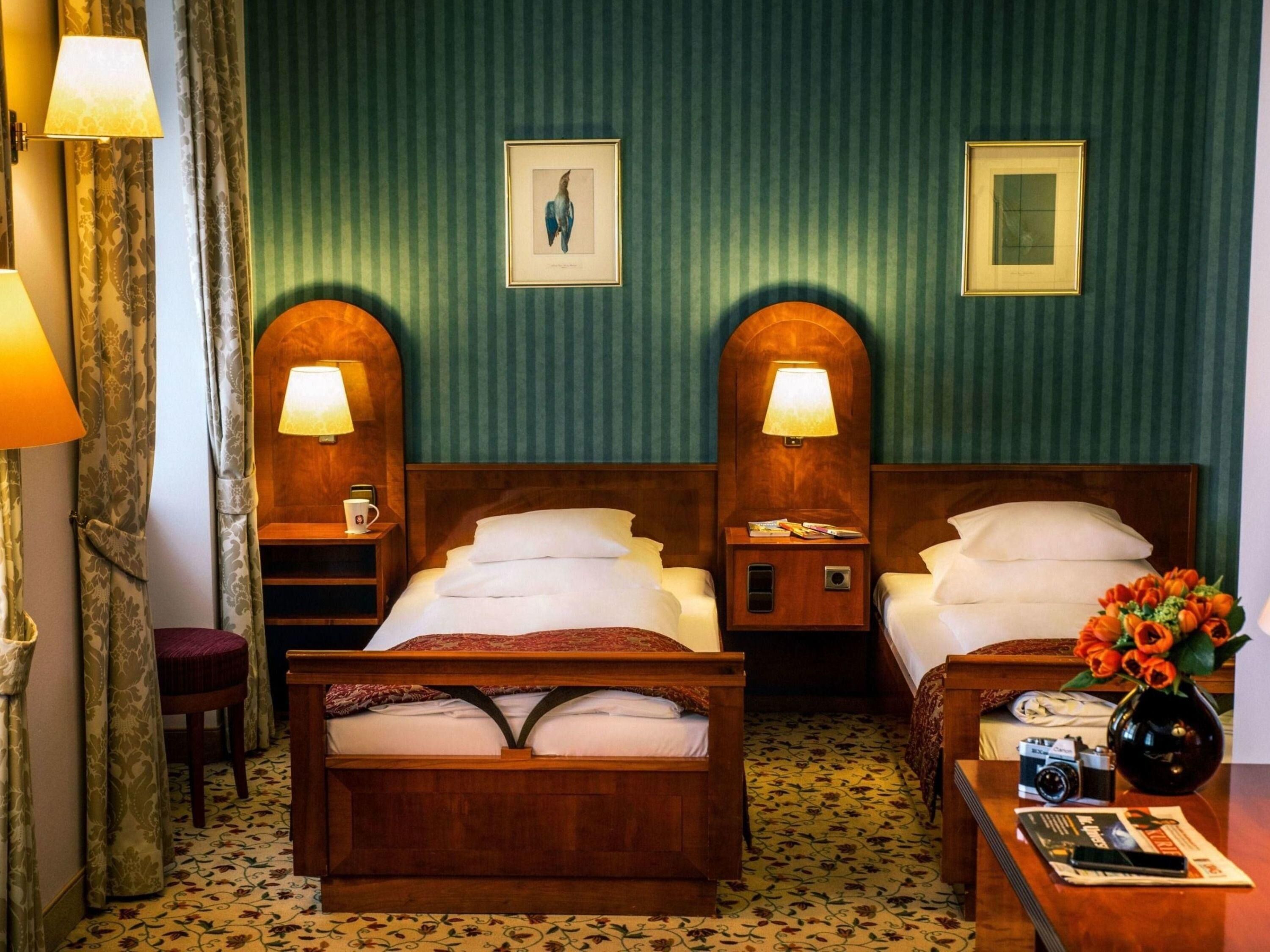 Quarto de hotel com duas camas, cabeceira de madeira, mesa de cabeceira com abajur, pequena mesa com um vaso de flores e um quadro na parede.
