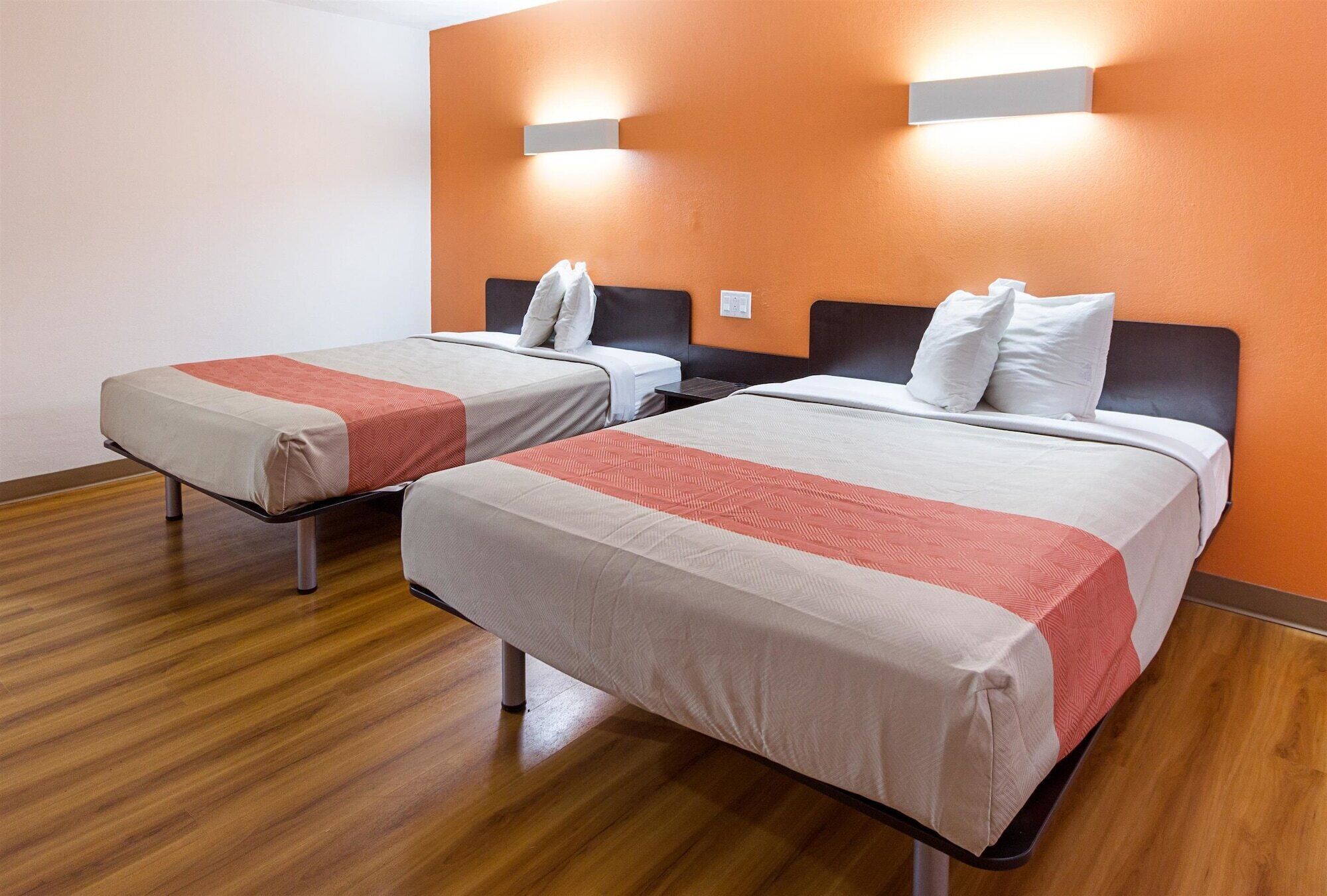 Dos camas individuales con ropa de cama blanca y roja, suelo de madera y paredes naranja con luces empotradas.
