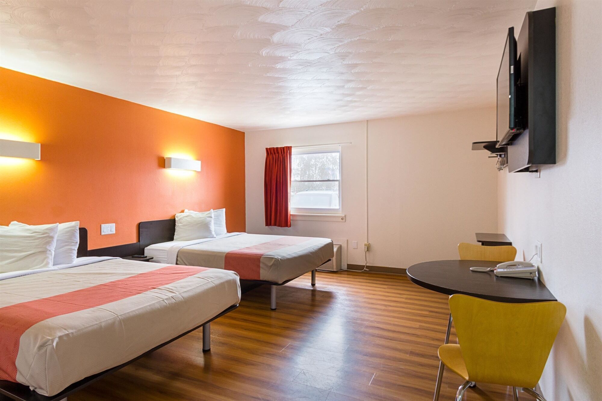 Una habitación de hotel con dos camas, una mesita pequeña, una silla amarilla, un televisor montado en la pared y un ventanal con cortinas rojas.