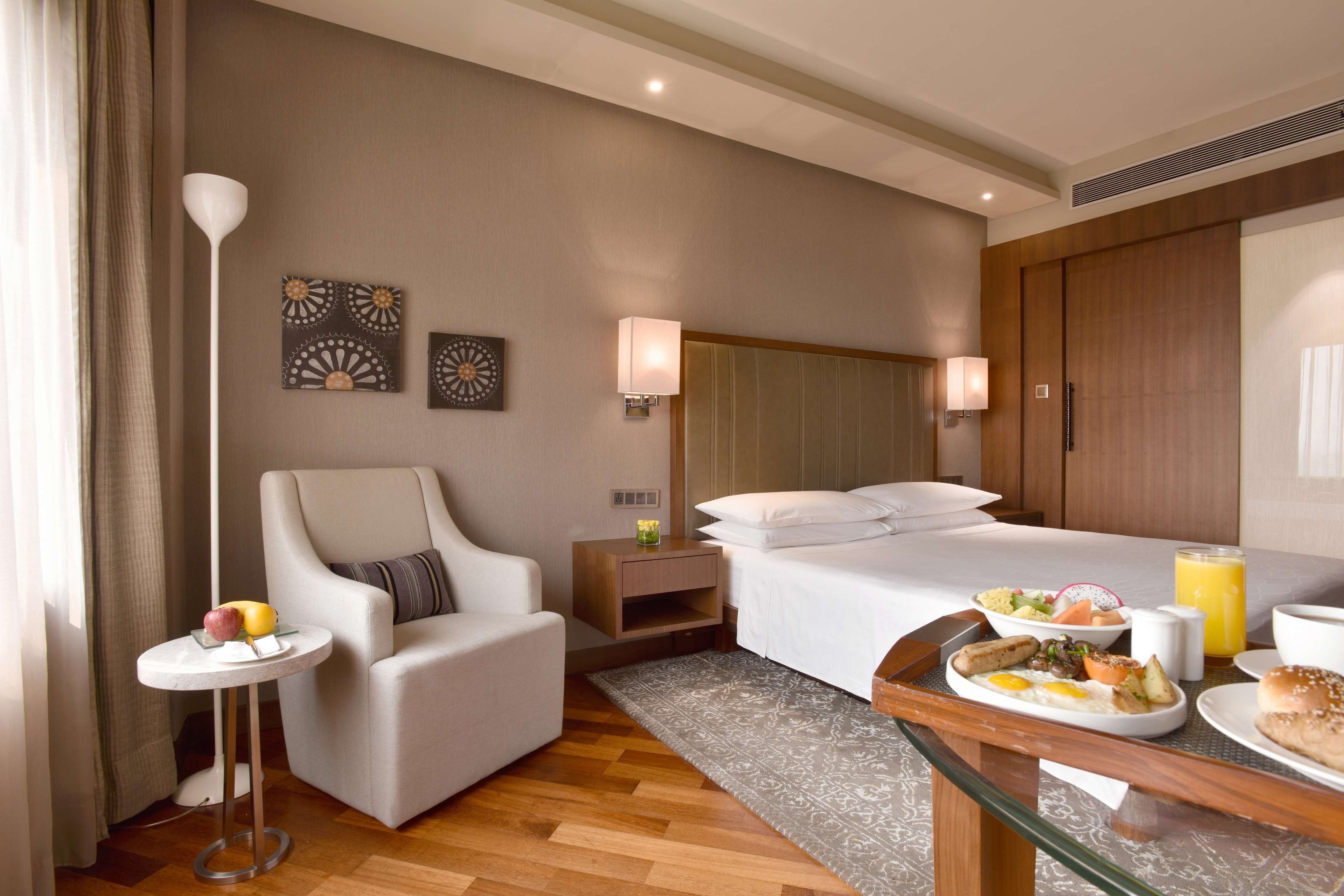 Ein Hotelzimmer mit Bett, Sessel, kleinem Tisch mit Obst und Frühstücksbrettern sowie einem Kunstwerk an der Wand.