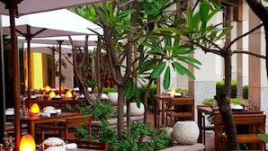 An outdoor dining area with wooden tables and chairs, surrounded by greenery and stone planters.