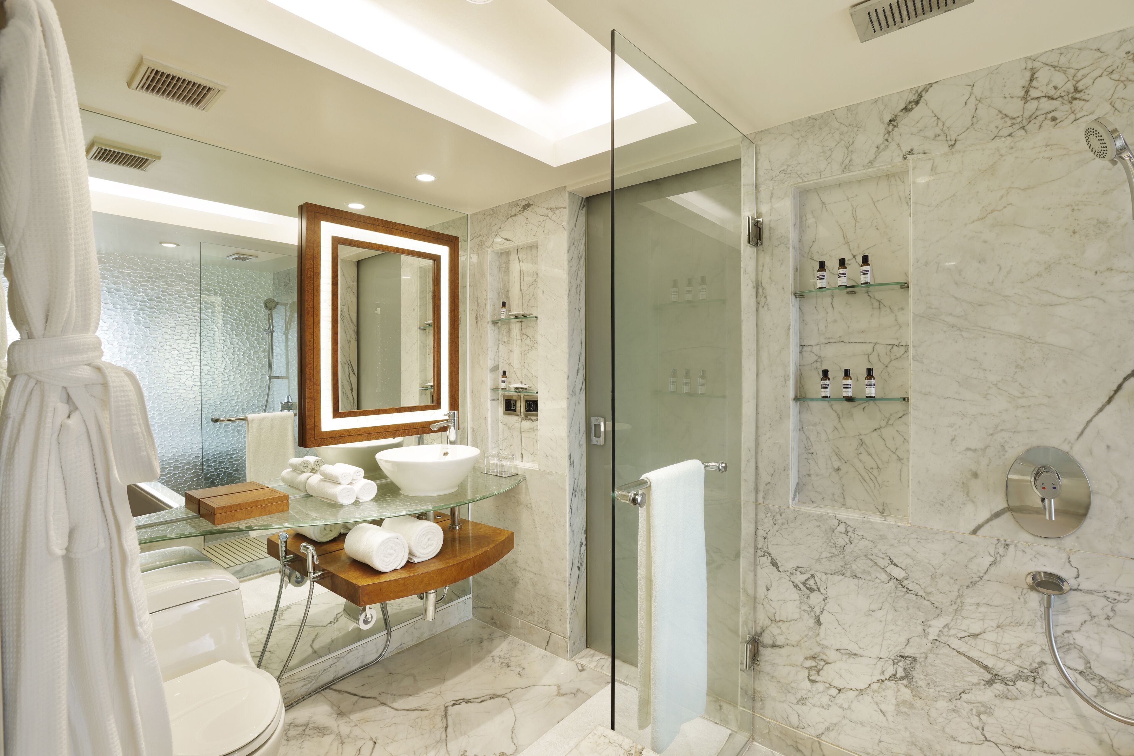 A modern bathroom with a glass shower enclosure, a marble-tiled floor, a glass sink, and a wooden vanity.