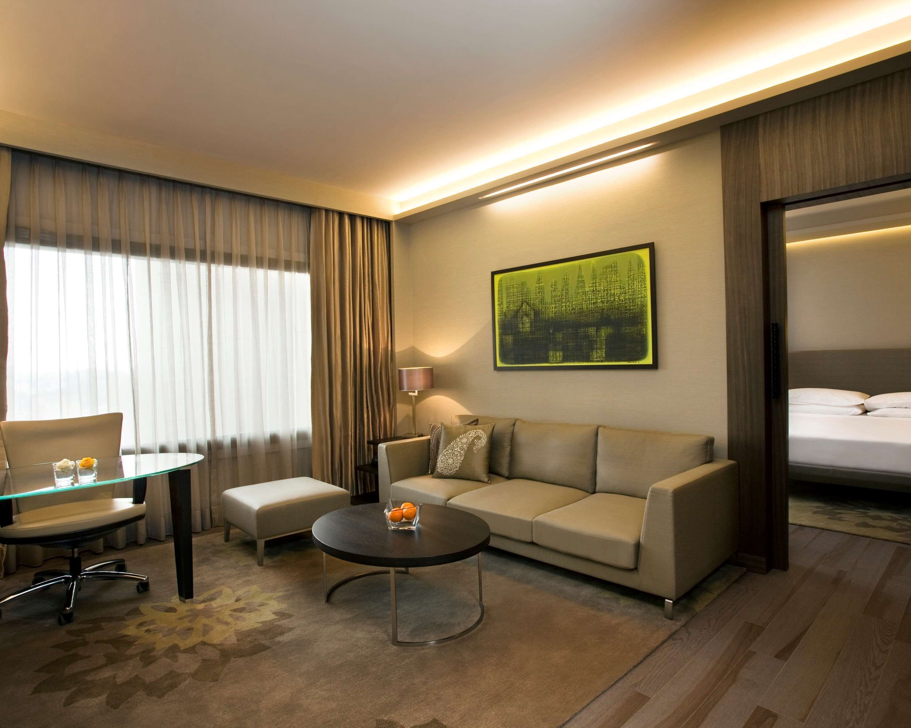 A hotel room with a sofa, a round coffee table, a chair, a bed, and a painting on the wall.