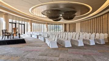 Un salón amplio con filas de sillas blancas, un escenario con sillas y ventanas grandes.