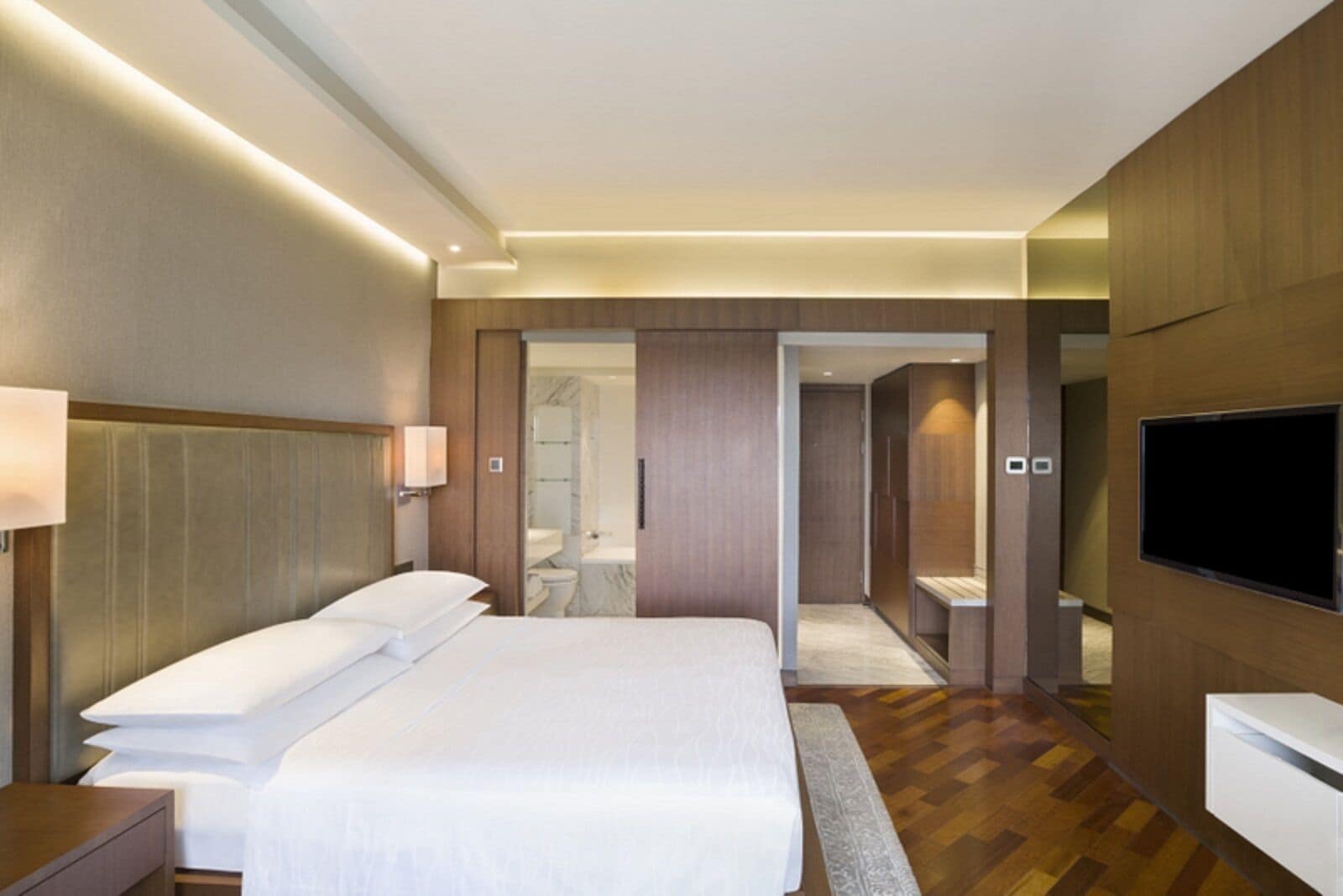 A modern hotel room with a large bed, a TV, a wardrobe, and a bathroom visible through an open door.
