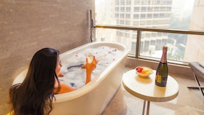 A woman in a bathtub with a bottle of champagne and a bowl of fruit on a small table beside her.