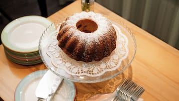 A bundt cake on a glass cake stand with a lace doily, surrounded by plates and cutlery on a wooden table.
