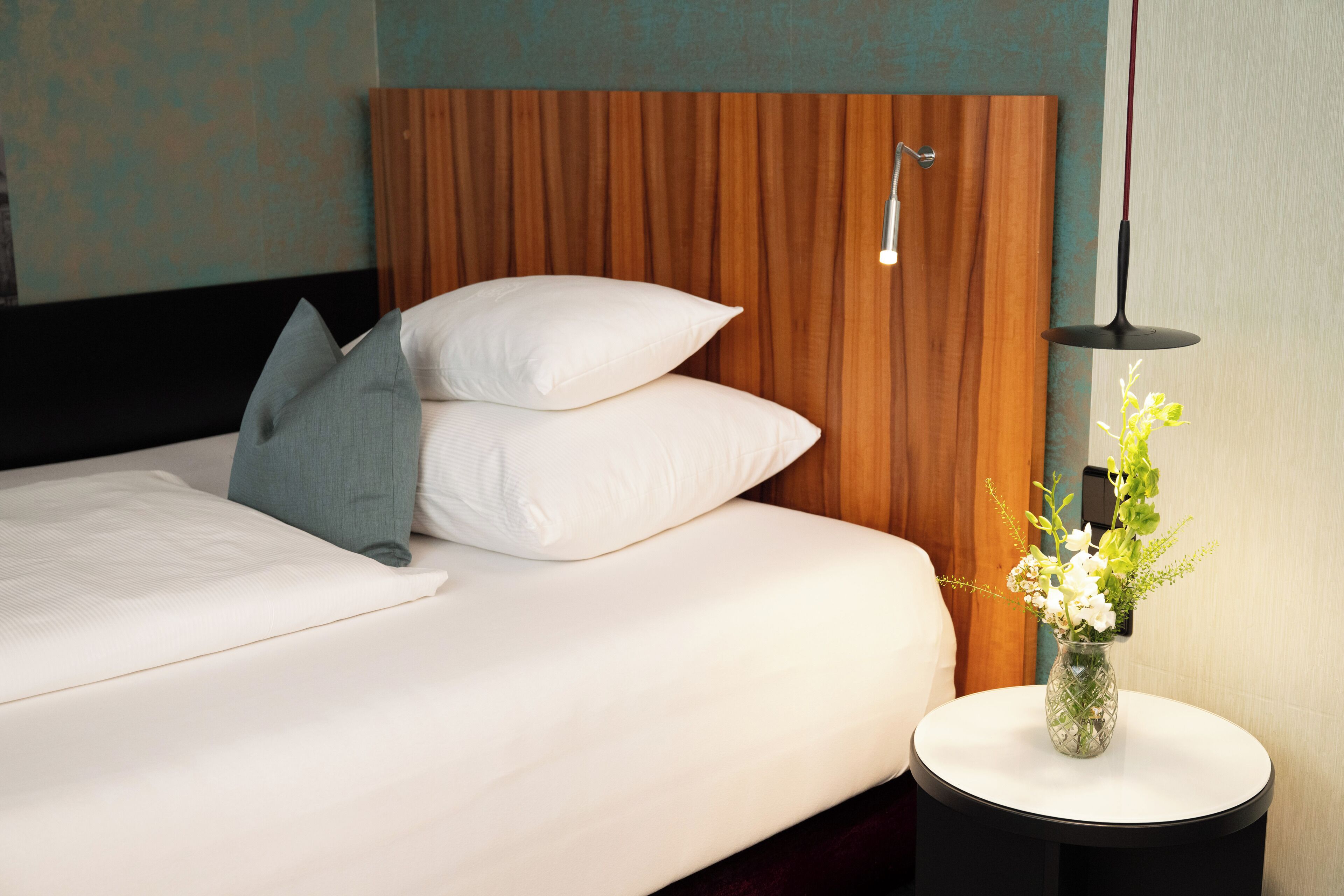 Una cama bien hecha con almohadas, un jarrón con flores en una mesita de noche y una lámpara moderna.