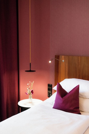 A hotel room with a wooden headboard, a white bed with a purple pillow, a small round table with a vase of flowers, and a hanging pendant light.