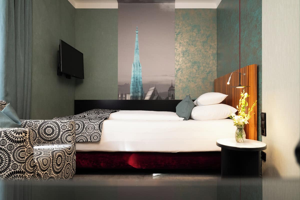 Quarto de hotel moderno com uma cama grande, televisão na parede, uma mesinha redonda com um vaso e um mural com vista para a cidade.