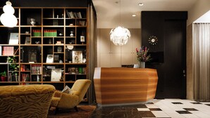 A reception desk with a modern design, a bookshelf filled with books and decorative items, a patterned armchair, and a checkered floor. - Hotel Capricorno (Vienna)