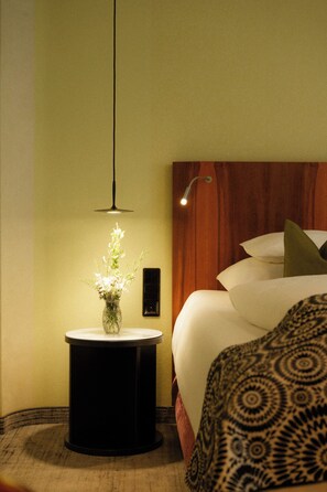 Camera d'albergo con un letto, un comodino con un vaso di fiori, una lampada a parete e una testiera in legno.