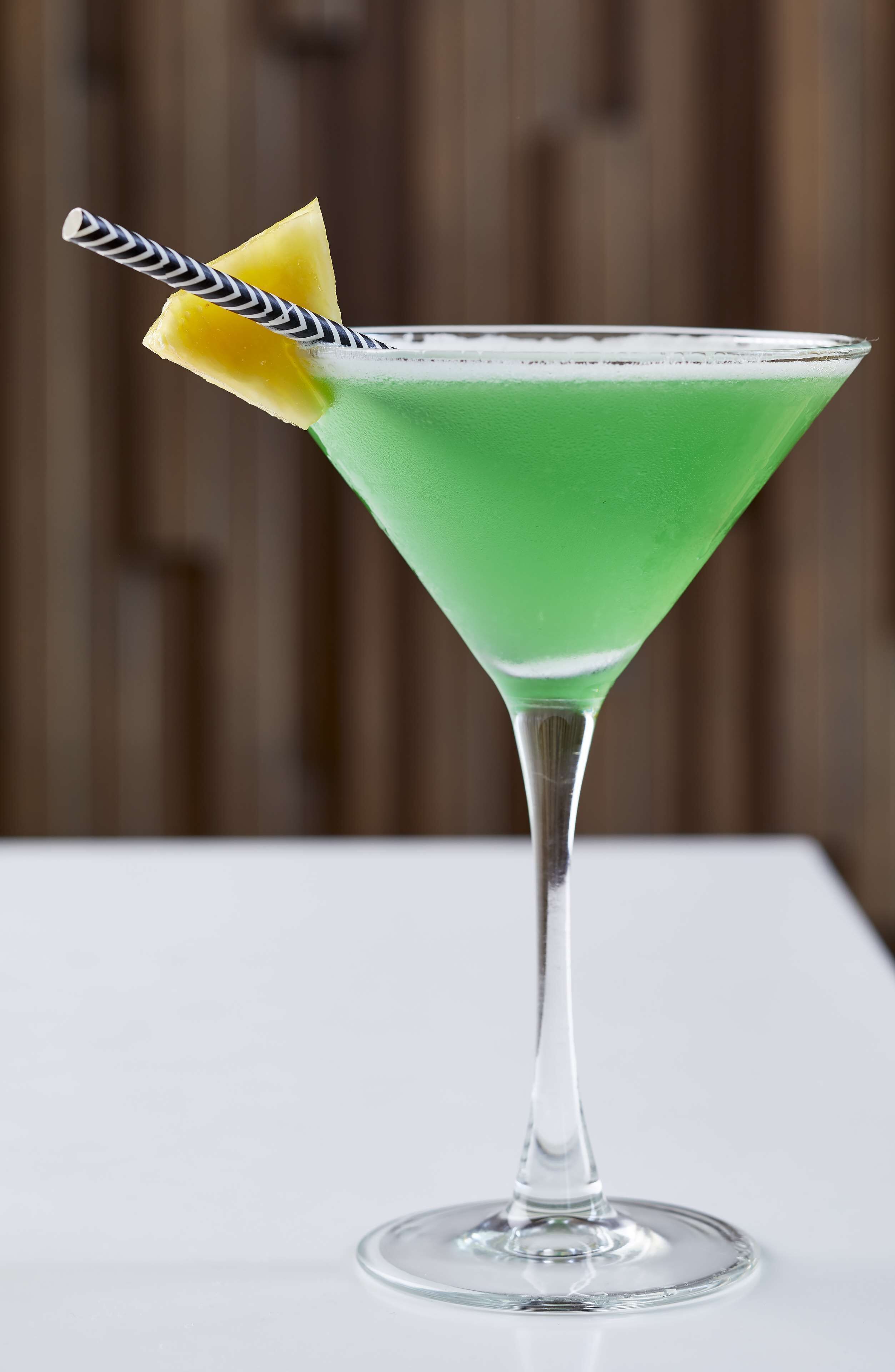 A martini glass with a green liquid, a yellow fruit garnish, and a striped straw.