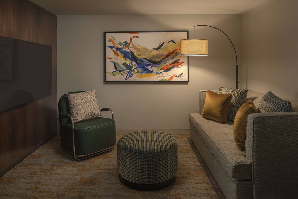 A modern living room with a green armchair, a patterned ottoman, a grey sofa with patterned cushions, and a large abstract painting on the wall.