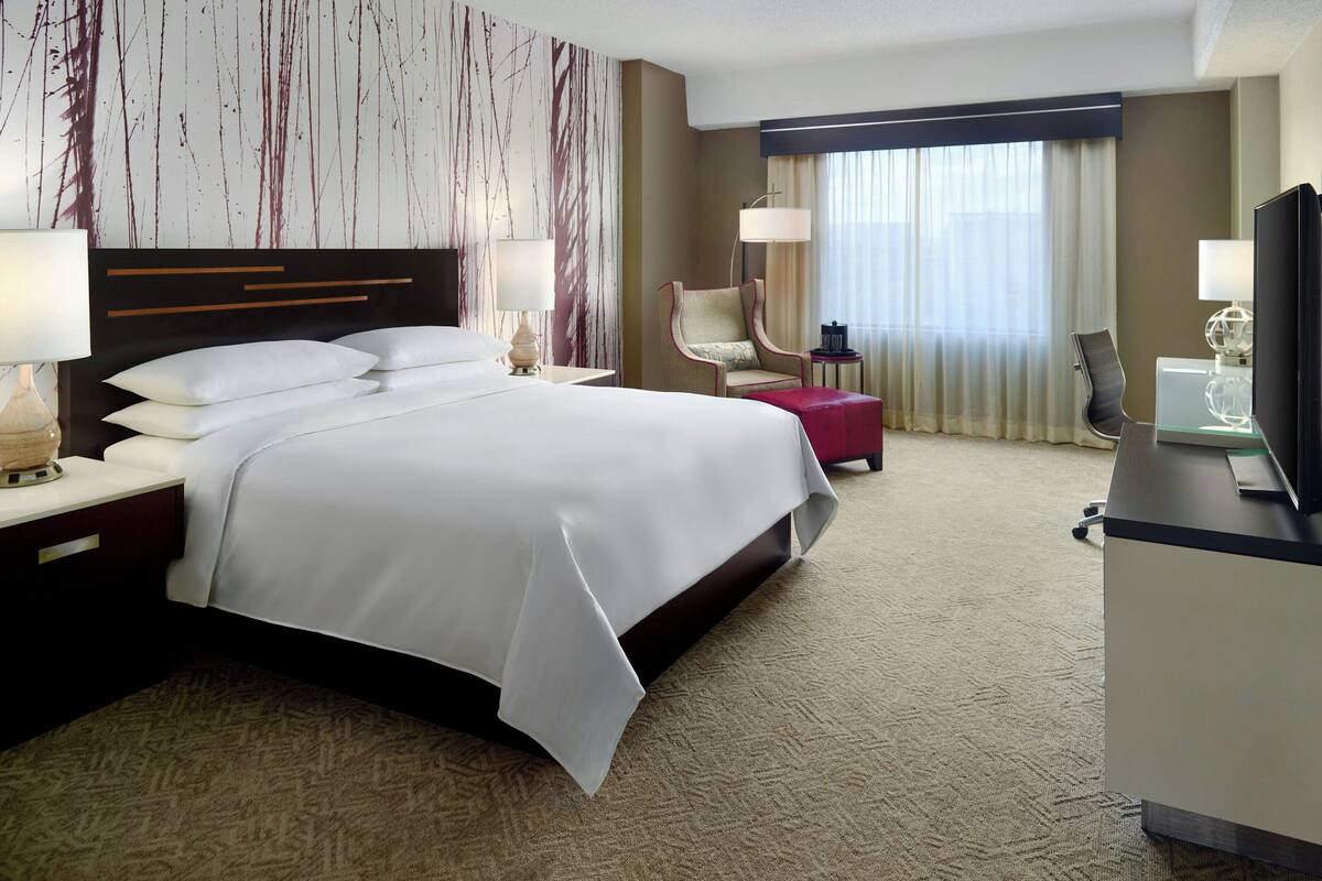 A hotel room with a large bed, a desk with a television, a chair, and a small table with a lamp.