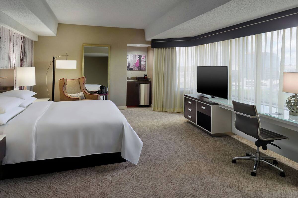 A hotel room with a bed, desk, chair, television, and a view of the city through large windows.