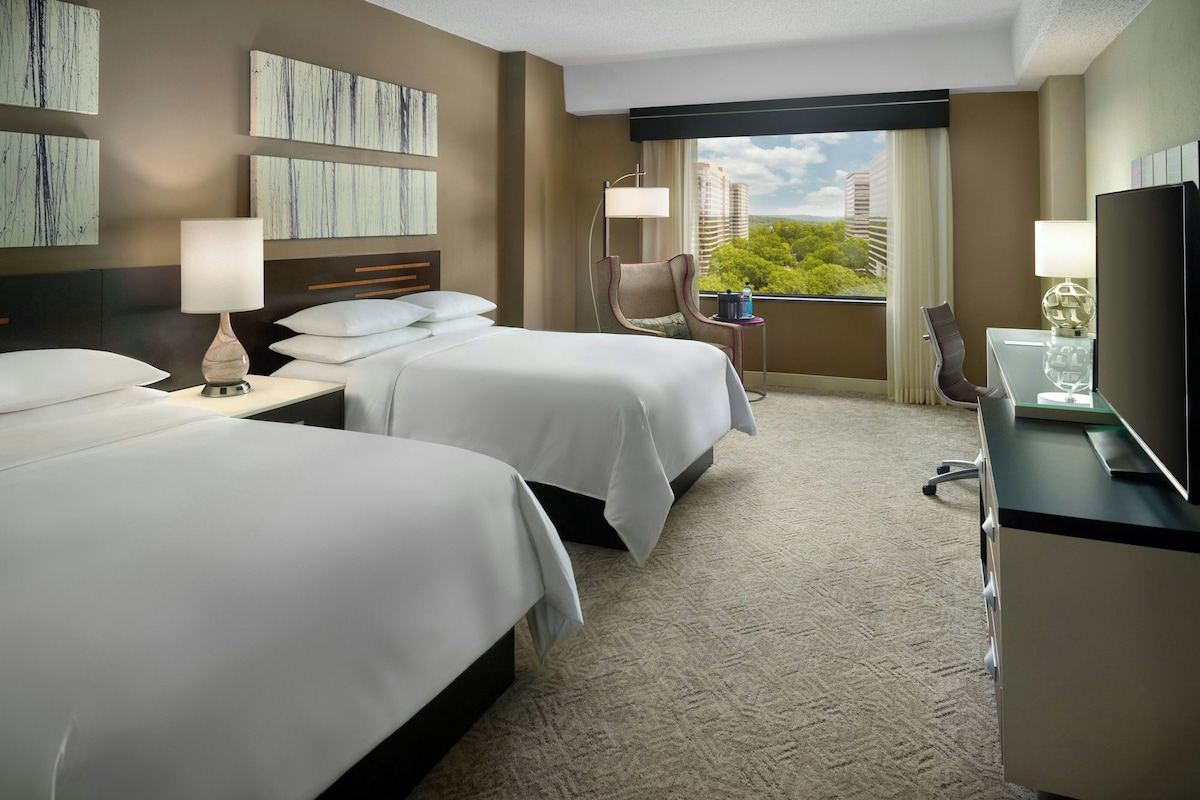 A hotel room with two beds, a desk, a chair, a TV, and a balcony with a city view.