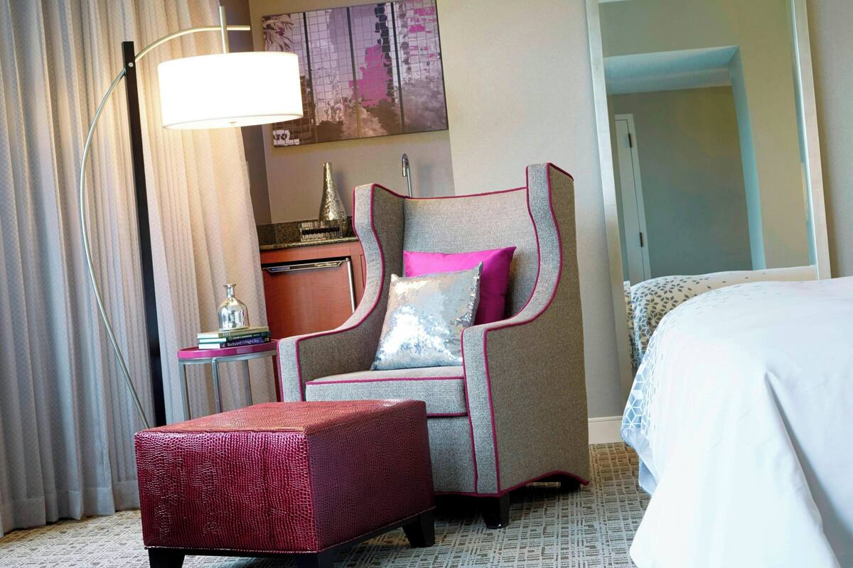 A hotel room with a bed, a chair with a cushion, a small table with a lamp, and a decorative item on the table.