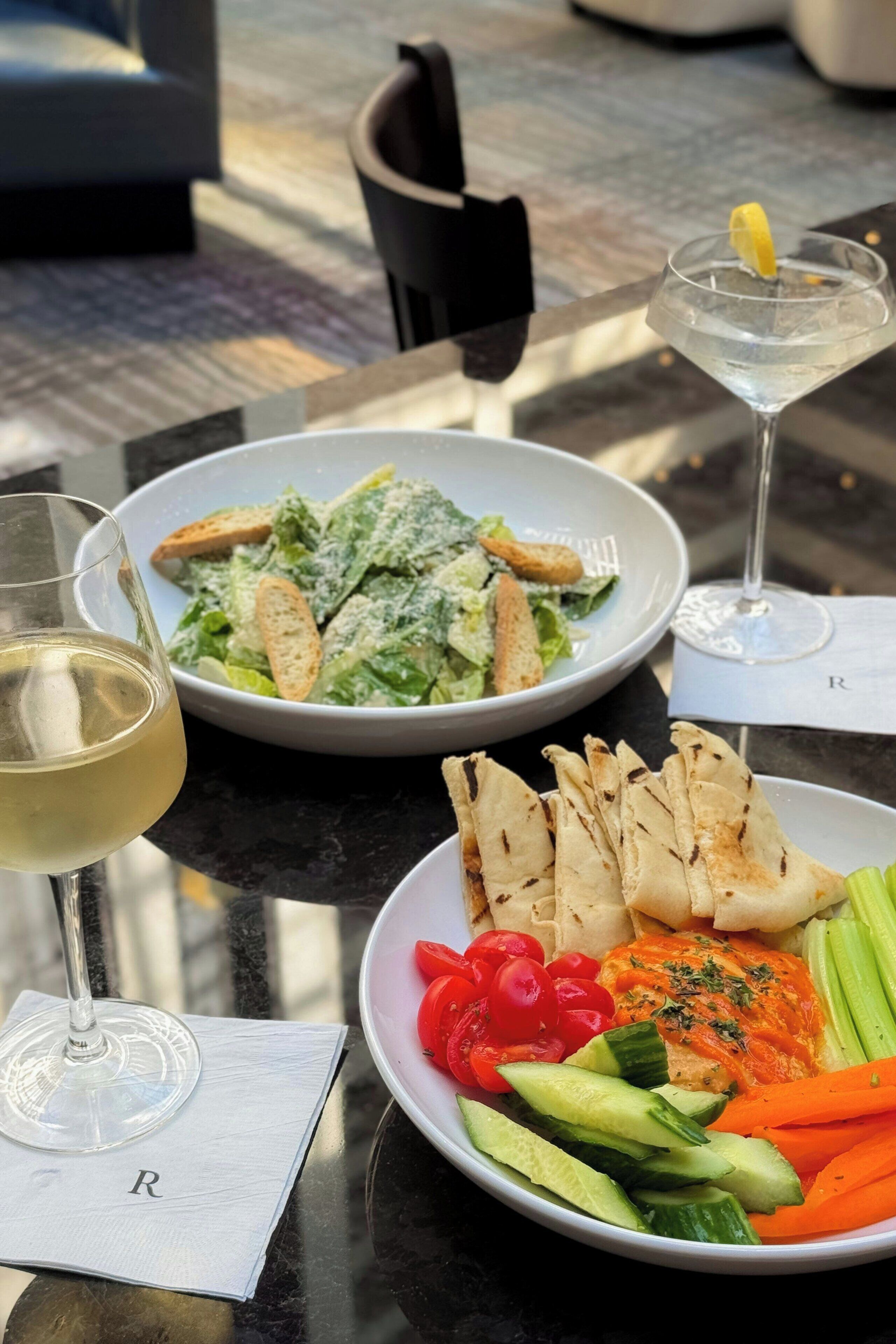 Et bord med en salat, en portion hummus med grøntsager, et glas hvidvin og et martini-glas med citron.