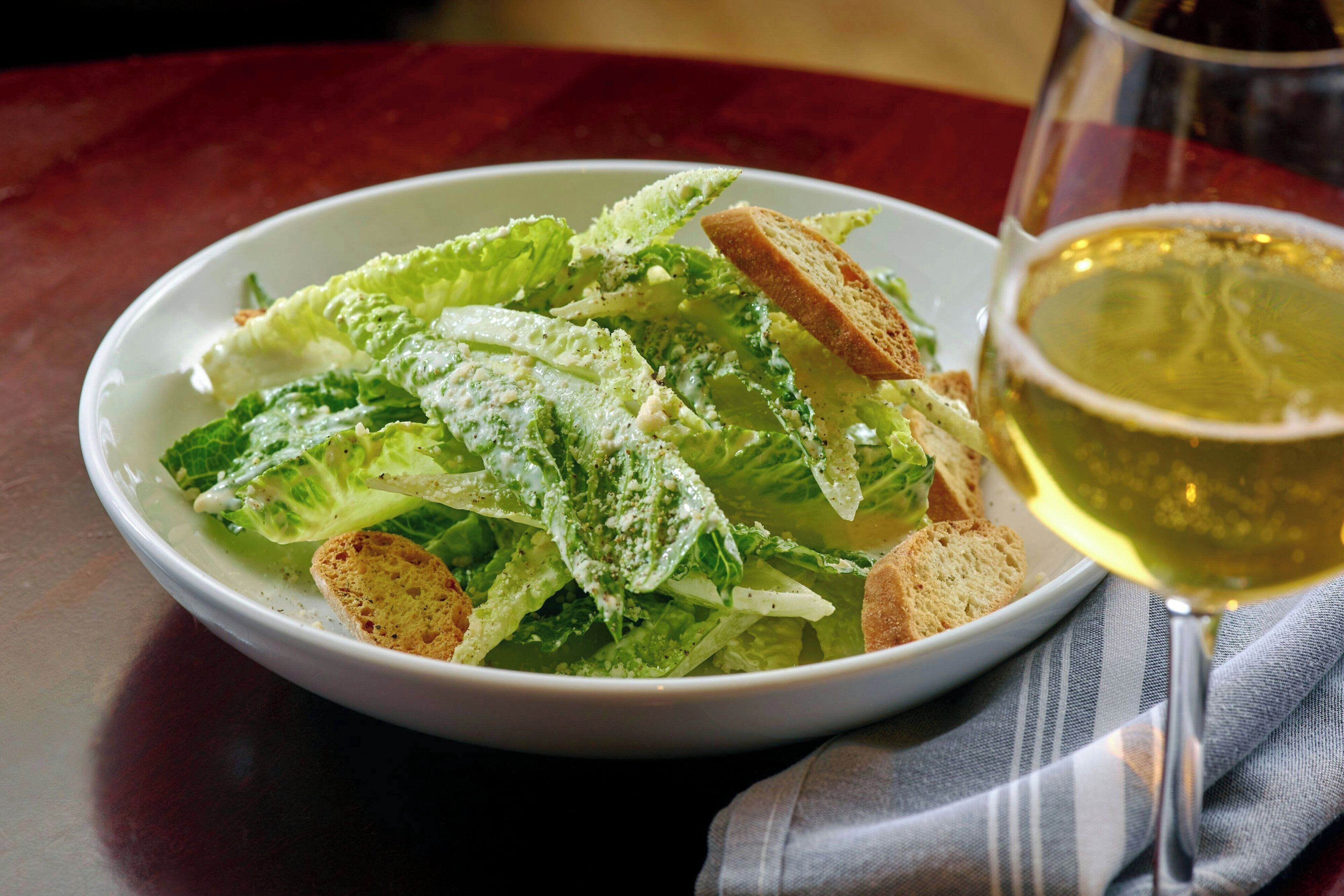 A bowl of salad with croutons and a glass of white wine on a table.