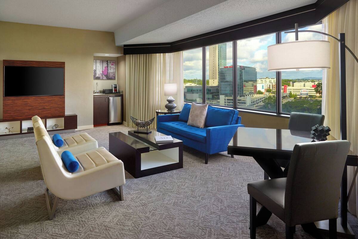 A hotel room with a large window, a flat-screen TV, a blue sofa, a white armchair, and a small table.
