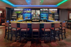 Zona de bar con barra central, diversas opciones de asientos y una pantalla grande mostrando un evento deportivo.