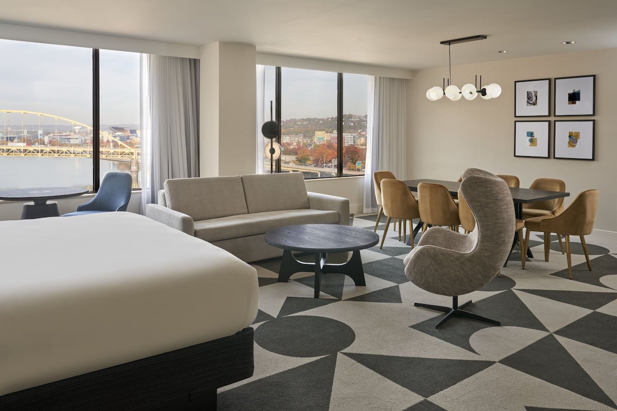 Habitación de hotel con una cama grande, un sofá, una mesa de comedor con sillas, una mesita de centro redonda y vistas a un puente y al paisaje urbano a través de grandes ventanales.