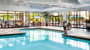 Una piscina interior con sillas de descanso y mesas, ventanas amplias y un techo con vigas a la vista.