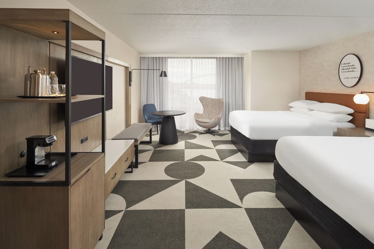 Habitación de hotel con cama, escritorio con cafetera, silla, mesita y ventana con cortinas.