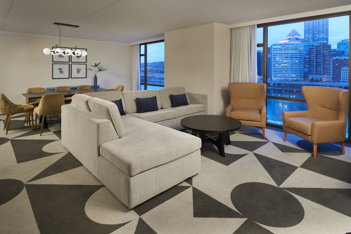 Una habitación de hotel moderna con un sofá grande, sillones, una mesa de comedor y vistas a la ciudad a través de amplias ventanas.